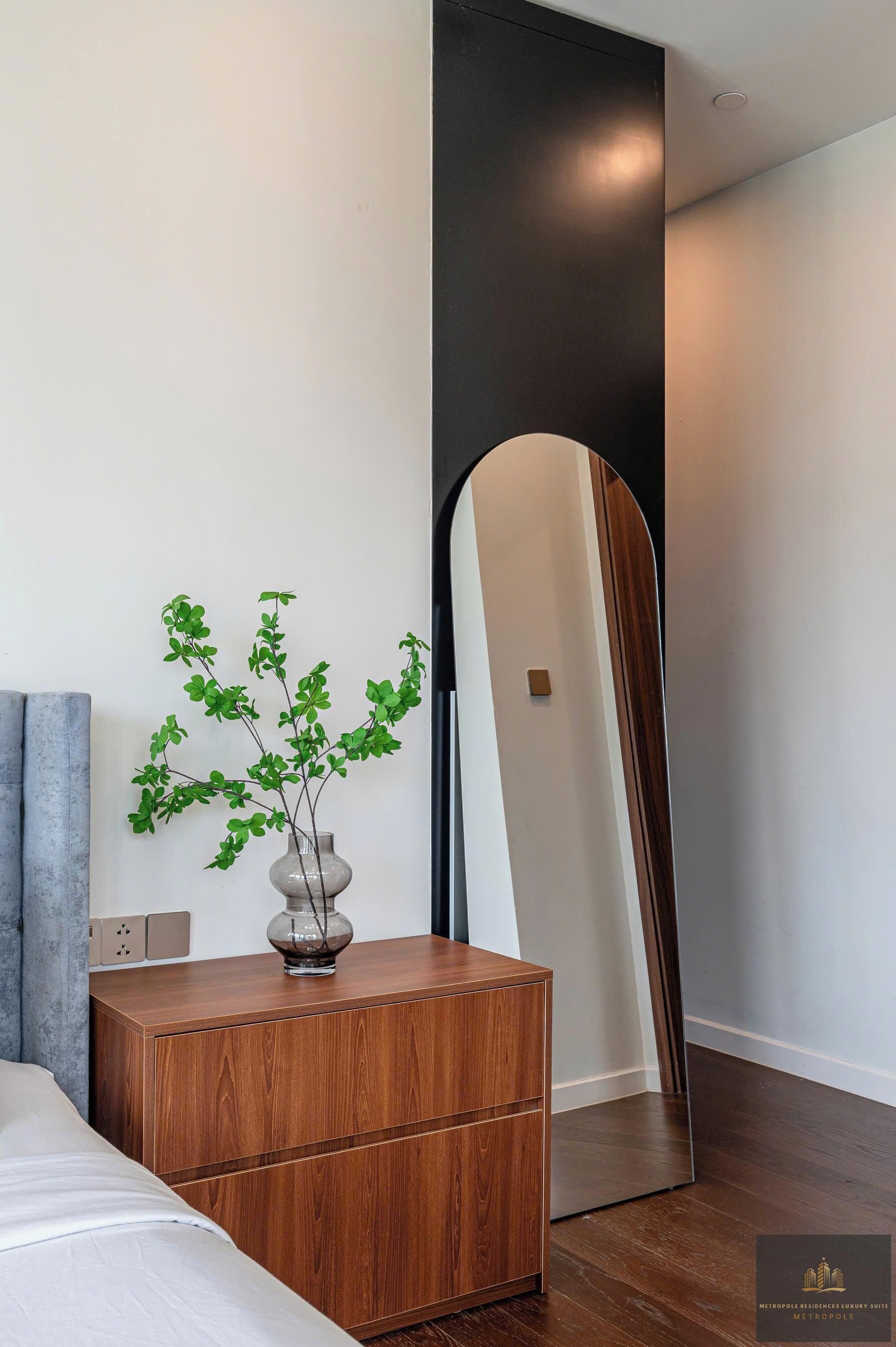 A modern bedroom with a wooden nightstand, a vase with green plants, a large mirror, and a bed with white bedding.