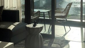 A modern living room with a large window overlooking a cityscape and a river. There are two wicker chairs and a small table. - Metropole Residences Luxury Suite (Ho Chi Minh City)