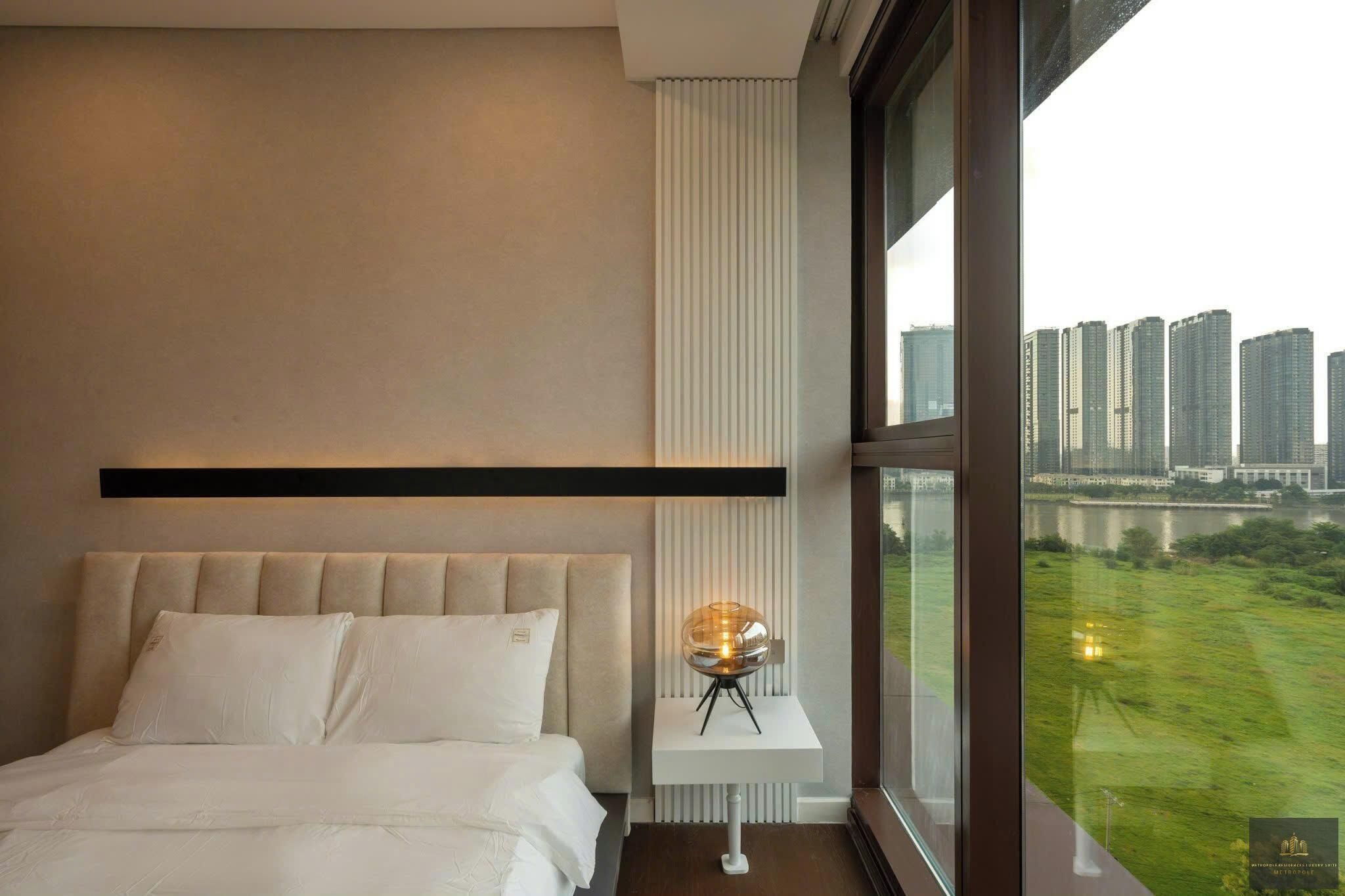 A modern hotel room with a large bed, a bedside table with a lamp, a view of high-rise buildings, and a cityscape.