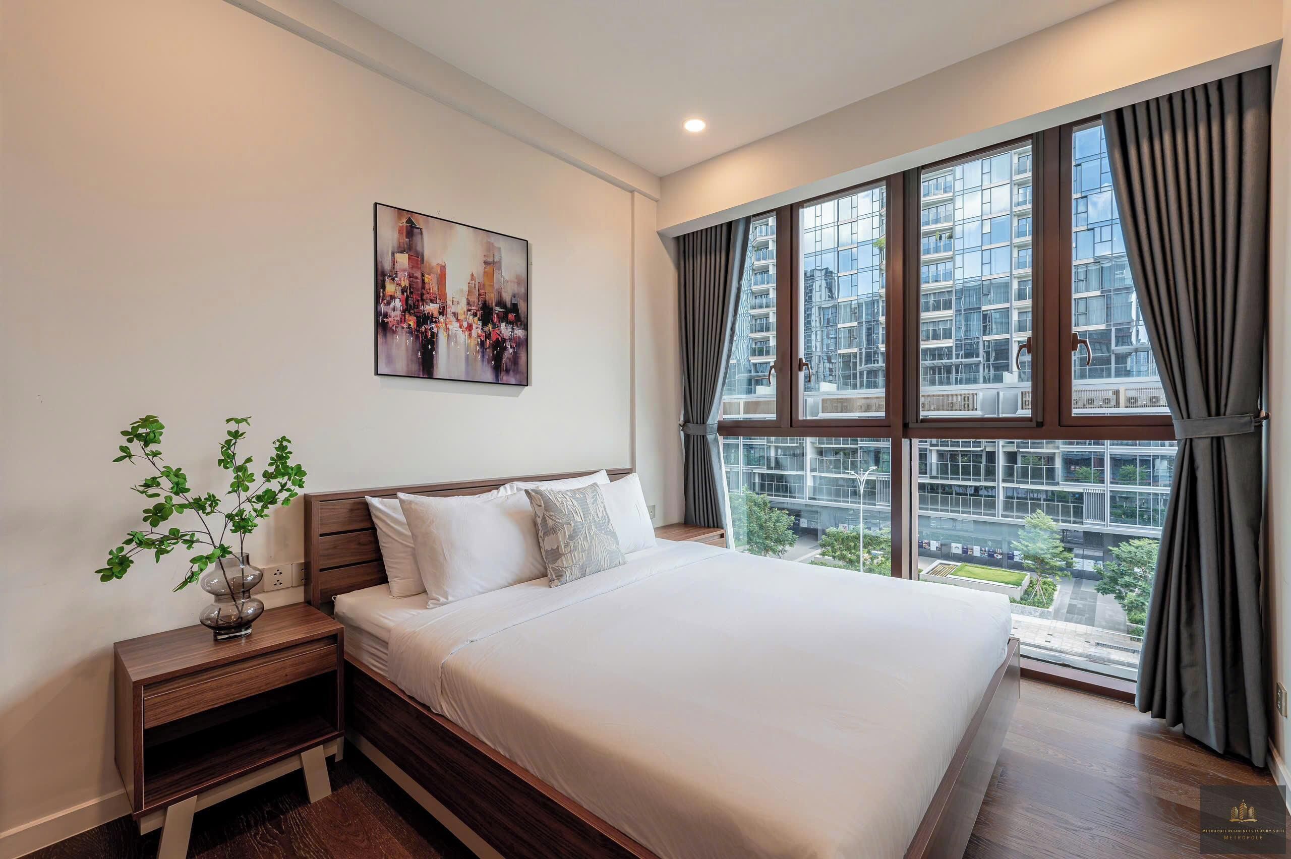 A modern hotel room with a large bed, a nightstand with a plant, a cityscape painting, and large windows with curtains.