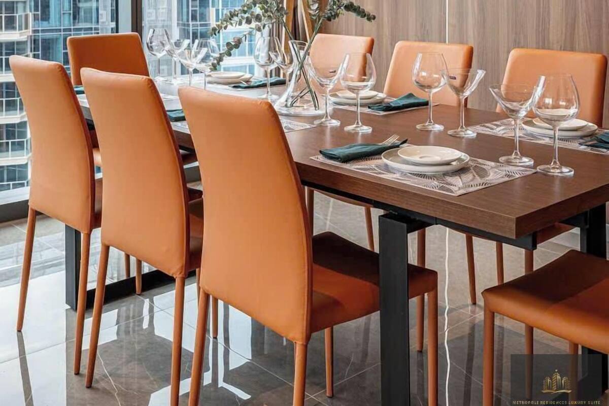 Ein moderner Essbereich mit einem großen Tisch für sechs Personen, umgeben von orangefarbenen Stühlen, einer dekorativen Kronleuchter und Blick auf eine Stadtkulisse durch große Fenster.