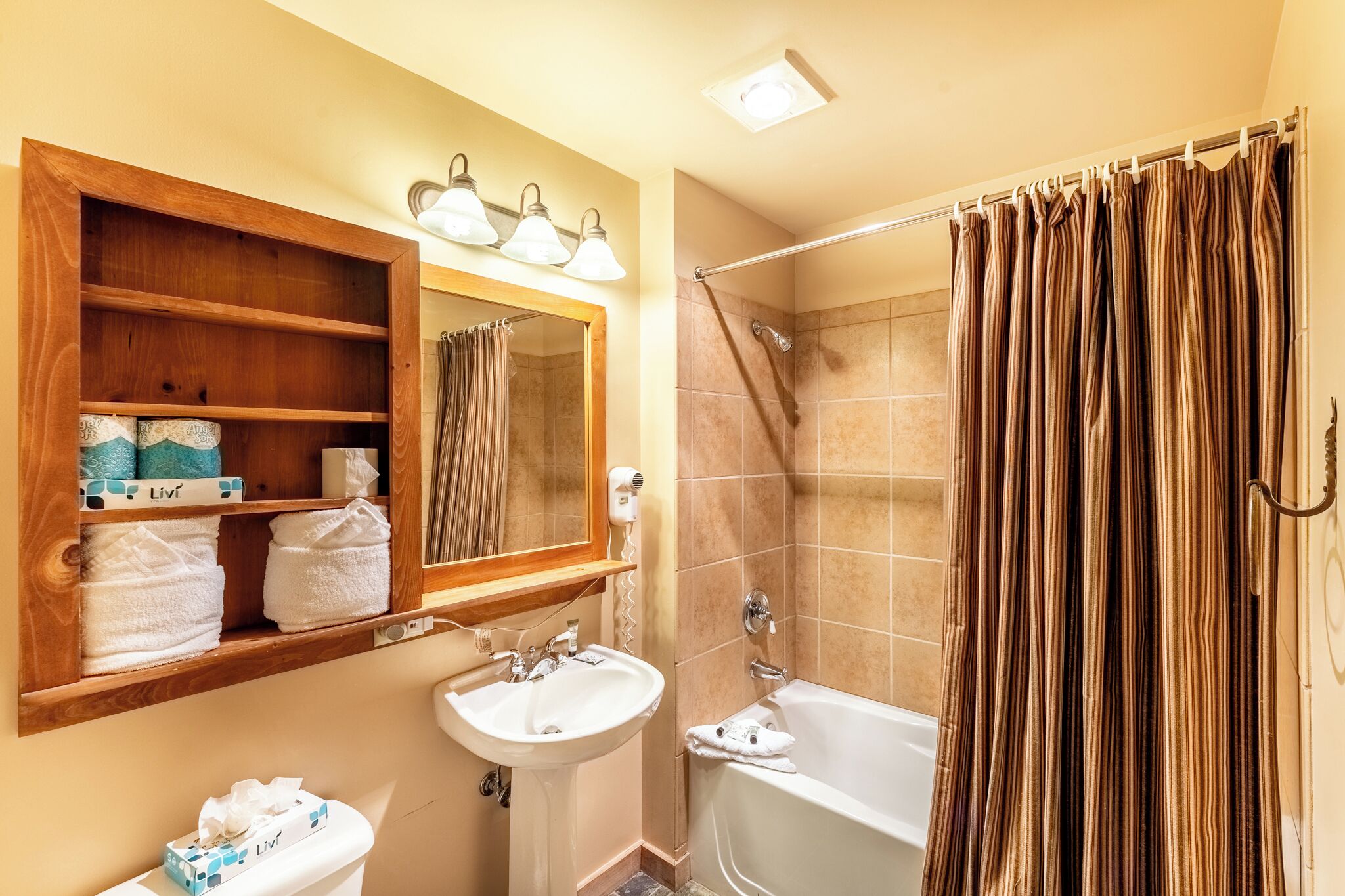 A bathroom with a wooden shelf holding towels, a sink, a mirror, and a shower curtain.