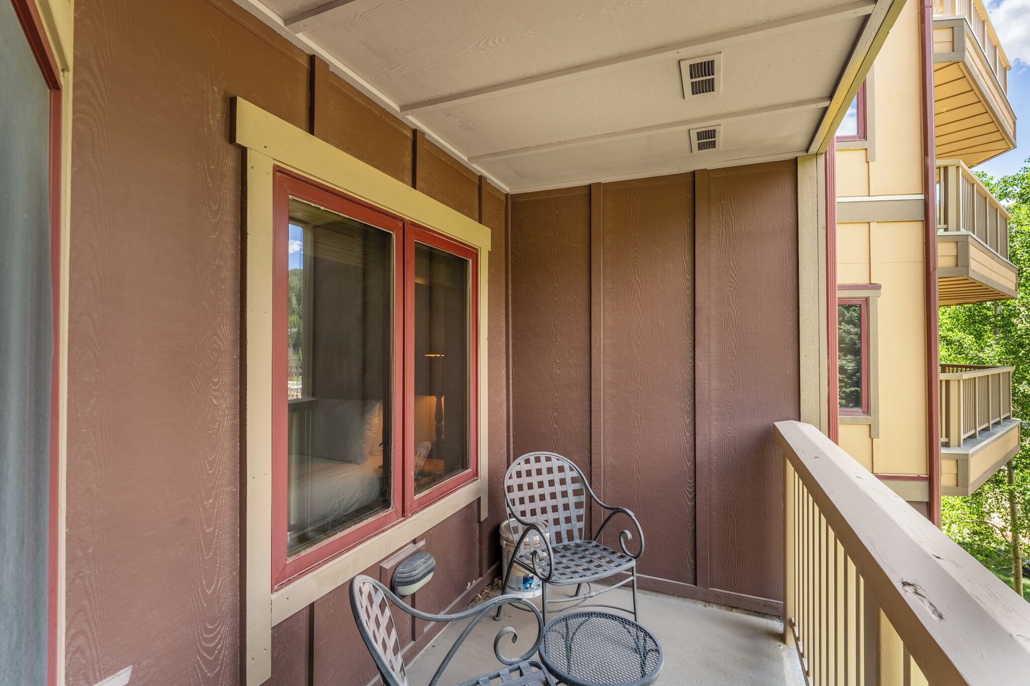 A balcony with two metal chairs, a window with a red frame, and a view of a tree.