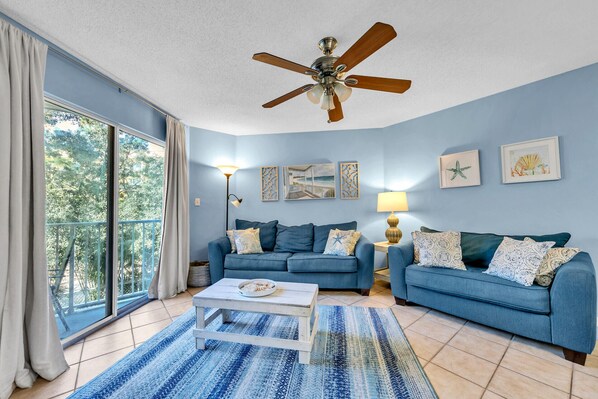 A living room with two blue sofas, a white coffee table, a ceiling fan, and a large window with curtains.