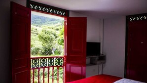 A room with a bed, a TV, and a balcony with red doors and a view of greenery.
