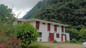 A white building with red doors and windows, surrounded by lush greenery and a hill in the background.