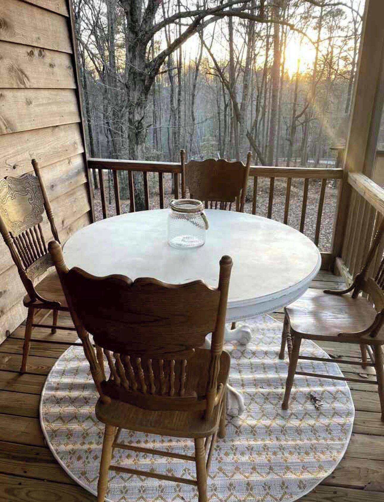 Una mesa redonda de madera con sillas en una terraza, rodeada por una baranda de madera, con vistas a árboles al fondo.