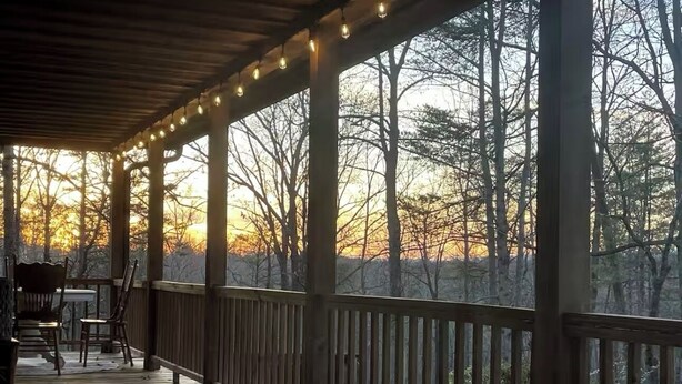 Terraza de madera con luces cálidas, sillas y vista a árboles al atardecer.