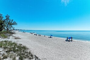 A wide sandy beach with a clear blue sky, some trees, and a few people and umbrellas. - The Magic Dolphin B, great Holmes Beach location, steps to gulf beaches (Holmes Beach)