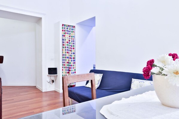 A room with a wooden floor, a blue sofa, a white vase with flowers, and a colorful curtain.