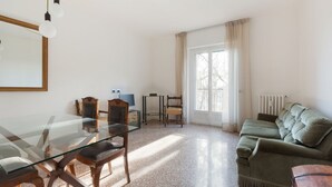 A spacious living area with a glass dining table, wooden chairs, a green sofa, and a large window providing natural light. - GuestHost - Comfortable 100sqm flat located on the fourth floor (with lift), equipped with kitchen, living area, two bathrooms and two bedrooms that can accommodate up to 4 people. A short walk from Lambrate and Udine stations, it is conveniently located (Milano)