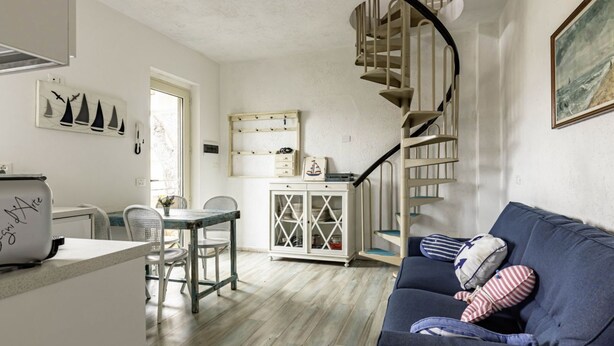 A cozy living room with a blue sofa, a dining area with a table and chairs, a staircase, and a kitchen area with cabinets and a refrigerator. - GuestHost - Pineta di Ponente Apartment (Viareggio)