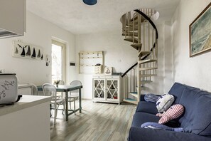 A cozy living room with a blue sofa, a dining area with a table and chairs, a staircase, and a kitchen area with cabinets and a refrigerator.