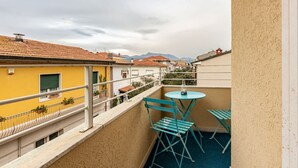 A balcony with a table and chairs, a railing, and a view of buildings and mountains. - GuestHost - Pineta di Ponente Apartment (Viareggio)