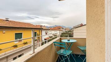Un balcón con mesa y sillas, barandilla y vistas a edificios y montañas.