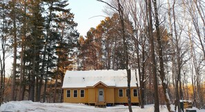 A wooden cabin with a snow-covered roof, surrounded by tall trees and a snowy ground.