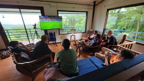 Un groupe de personnes est assis sur un balcon, devant un grand écran de télévision qui diffuse un événement sportif.
