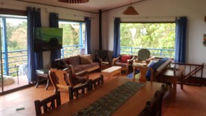 Un salon spacieux comprenant un canapé, des fauteuils, une table basse et un téléviseur. De grandes fenêtres sont agrémentées de rideaux, et il y a un balcon ainsi qu’un escalier menant à un autre étage.