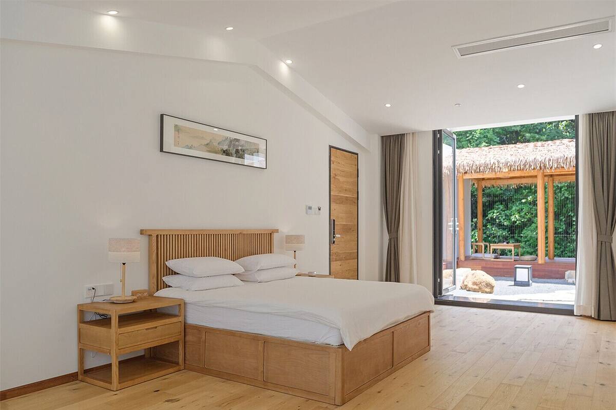A bedroom with a wooden bed, bedside tables, a nightstand, a large window with curtains, and a view of an outdoor area.