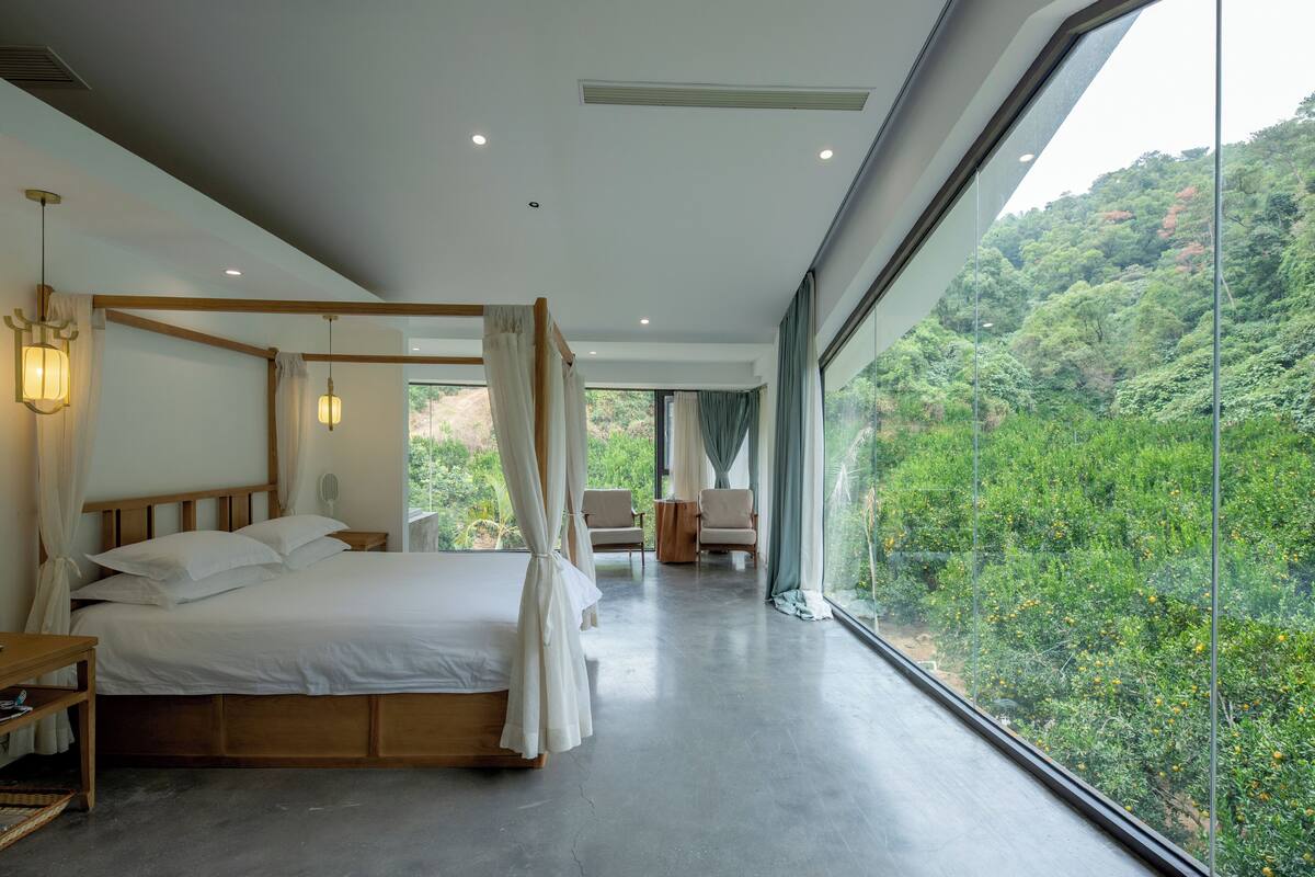 A spacious bedroom with a four-poster bed, a sitting area with chairs, and a large window offering a view of a lush forest.