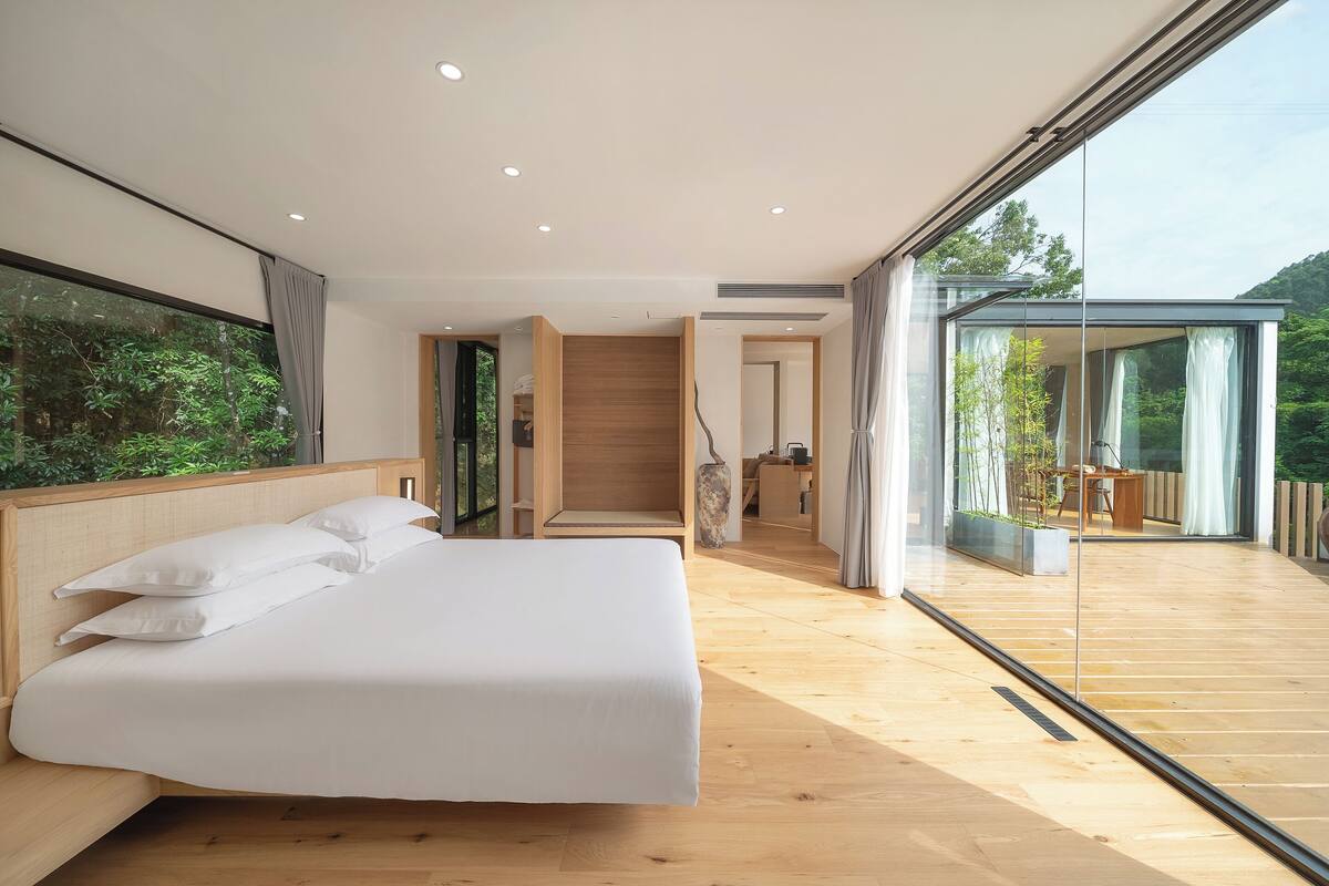 A modern bedroom with a large bed, wooden flooring, and a glass wall leading to a balcony with a view of greenery.