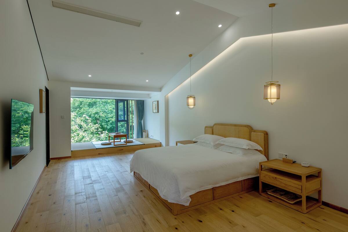 A modern bedroom with a large bed, bedside tables, and a view of greenery through the window.