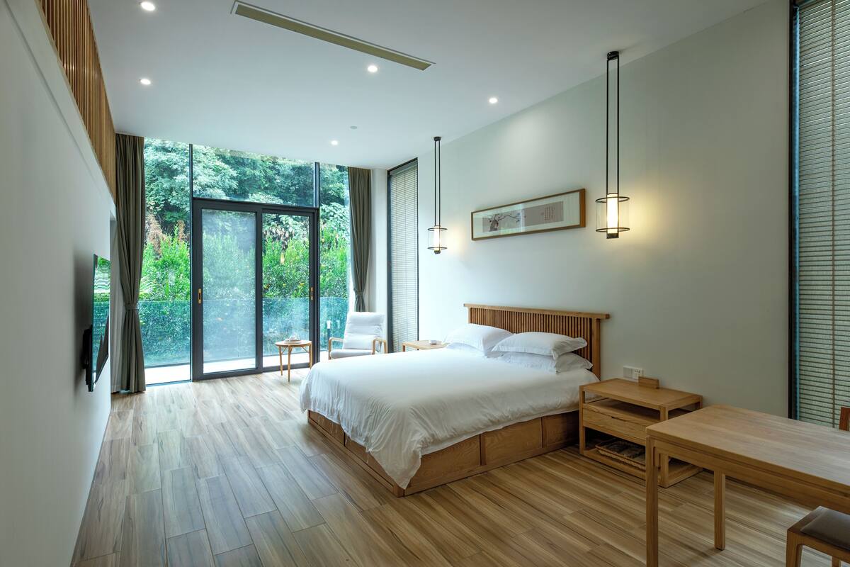 A modern bedroom with a large bed, wooden flooring, a desk, and a view of greenery through the window.