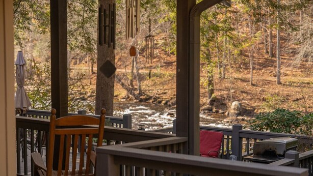 A wooden deck with a chair and a grill, overlooking a forested area with a stream.