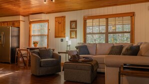 A cozy living room with a wooden ceiling, a large window with blinds, a sofa, a chair, and a dining table with chairs.