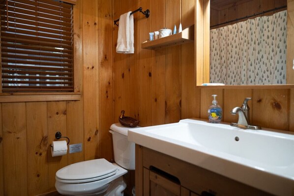 A wooden-paneled bathroom with a toilet, sink, and window with blinds. - River Zen, A Toccoa River Retreat (Blue Ridge)