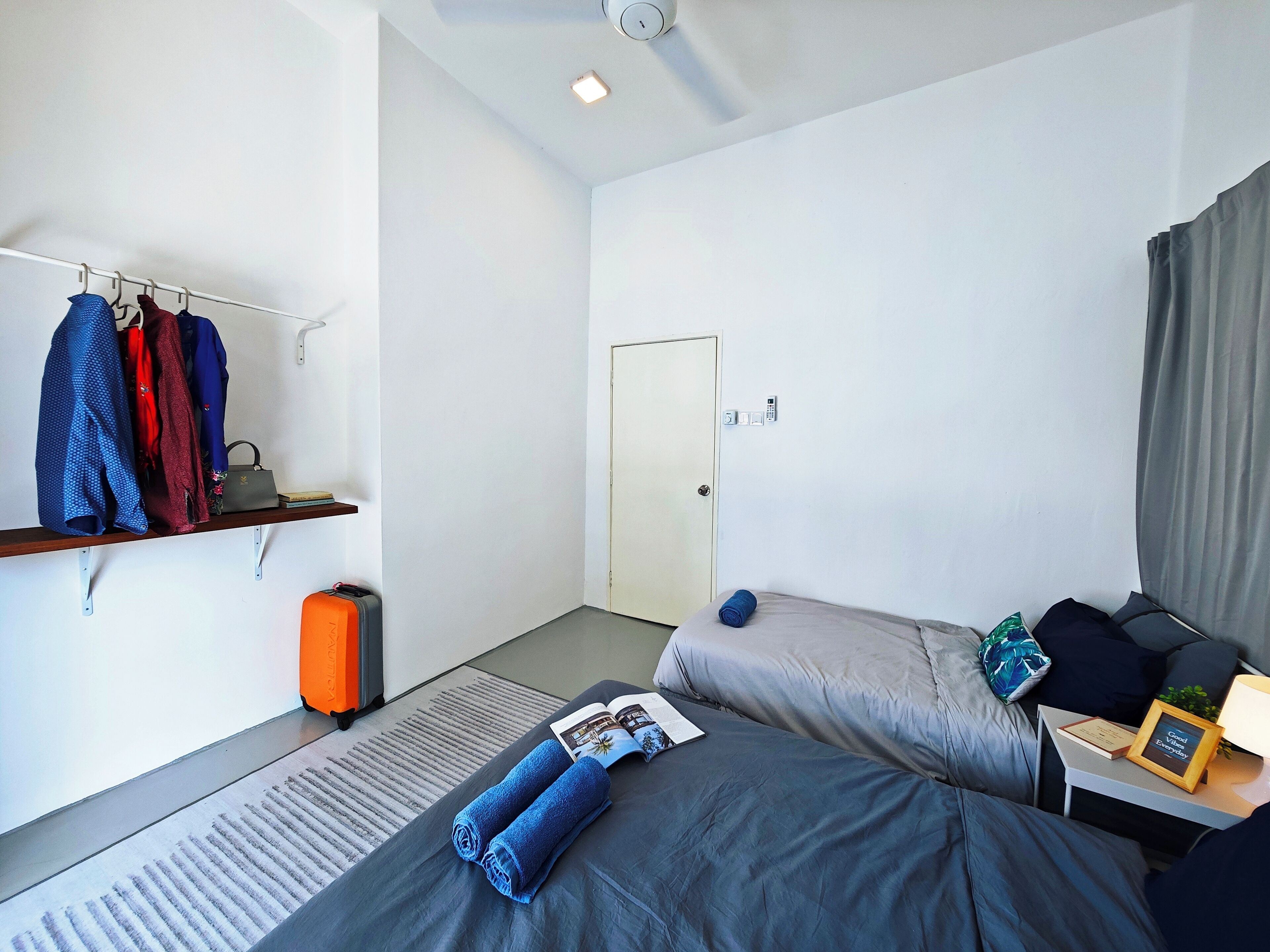 A hotel room with two beds, a small table with a lamp, a suitcase, and a shelf with clothes and a handbag.