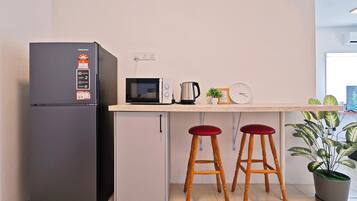 A compact kitchen with a refrigerator, microwave, kettle, and two stools.
