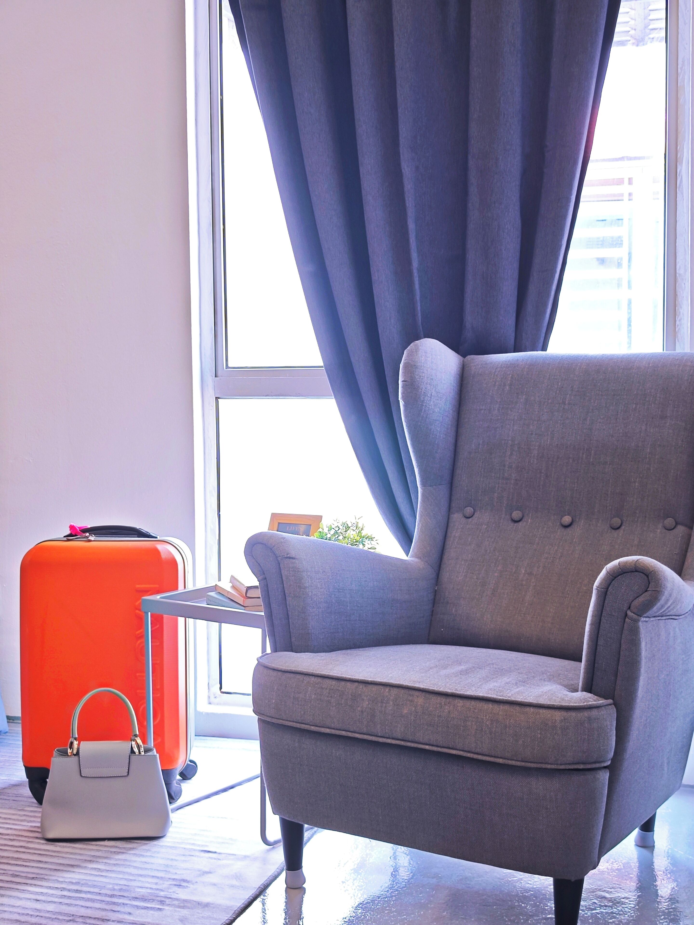 A grey armchair by a window with a view of a building, a red suitcase, and a white handbag on the floor.