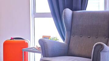 A grey armchair by a window with a view of a building, a red suitcase, and a white handbag on the floor.