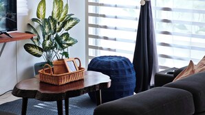 A modern living room with a dark sofa, a small table with a lamp, a potted plant, and a TV on a wooden stand. - Cosy Batu Kawan Homestay 12 pax ~ 5 mins to Design Village & IKEA ~ Entire Home (Simpang Ampat)