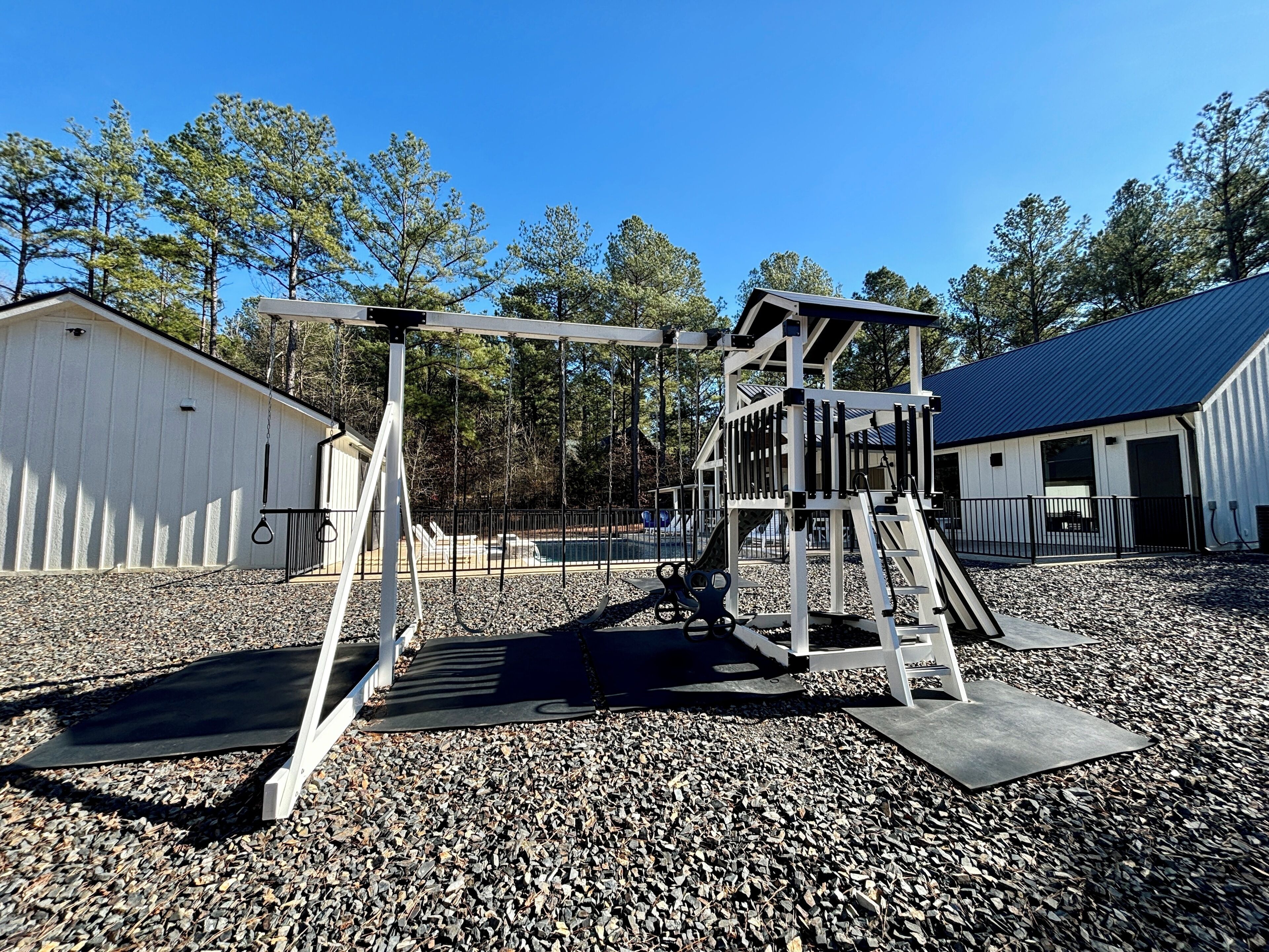 A playground with swings and climbing structures, surrounded by gravel and trees.