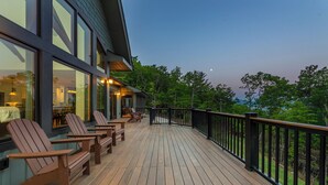 A wooden deck with Adirondack chairs and a glass railing overlooking a forested area.