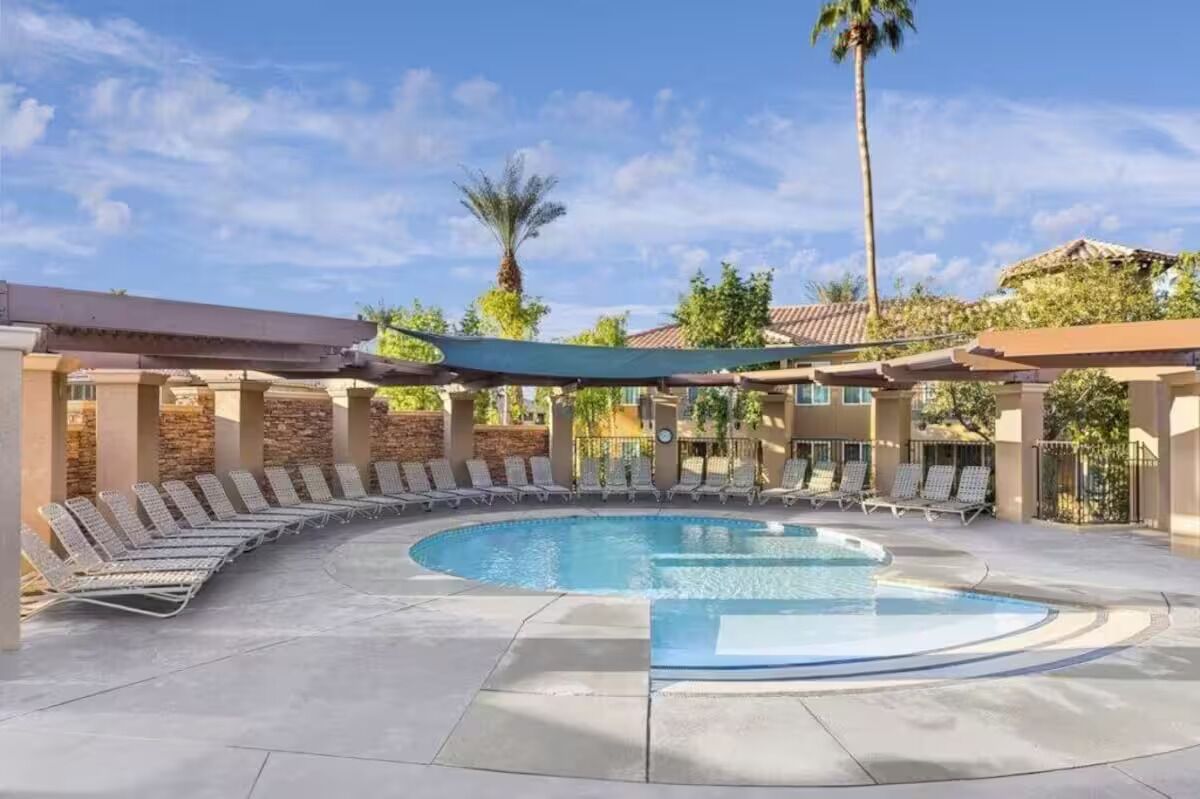 A circular pool with lounge chairs around it, shaded by a canopy, and surrounded by a building with a tiled roof and stone walls.