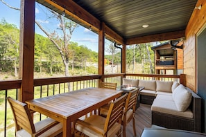 Una terraza techada al aire libre con una mesa y sillas de madera, un sofá de mimbre con cojines y vistas al bosque circundante.