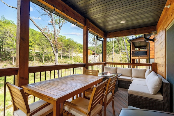 Eine überdachte Terrasse im Freien mit Holztisch und -stühlen, einer Rattan-Couch mit Kissen und Blick auf den umliegenden Wald.