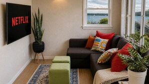 A living room with a sofa, ottomans, a potted plant, a TV, and a window with a view.