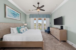 A bedroom with a bed, a ceiling fan, a TV, a dresser, and a view of the outdoors.