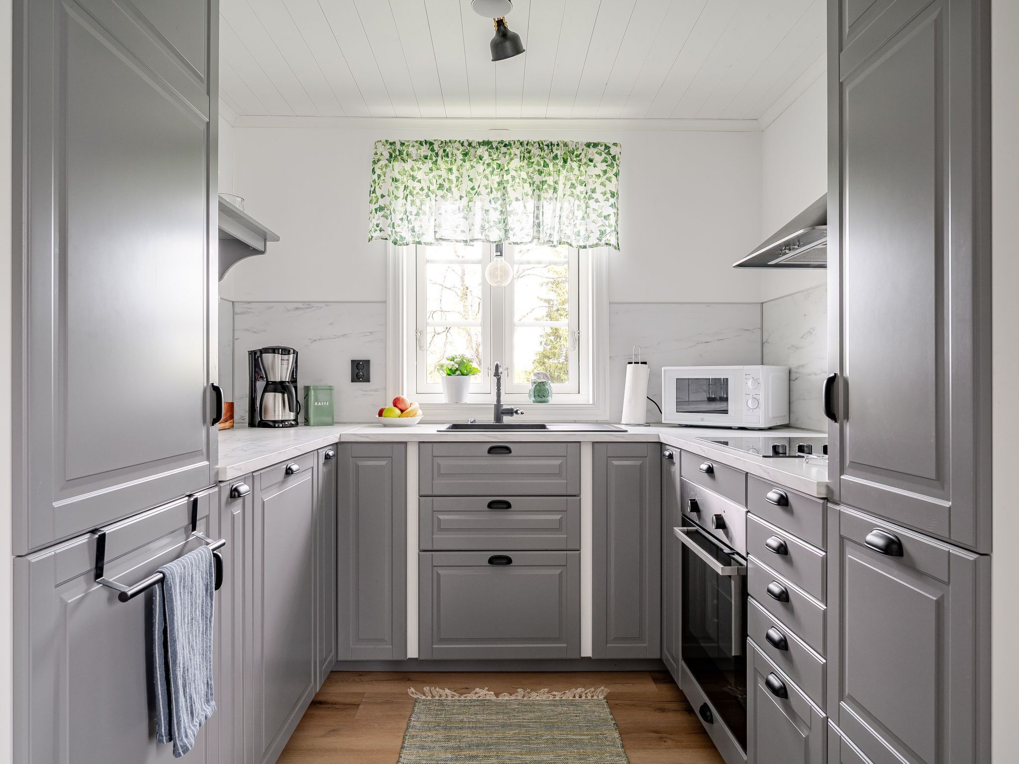 A modern kitchen with grey cabinets, a white countertop, a built-in microwave, and a window with a green floral curtain.
