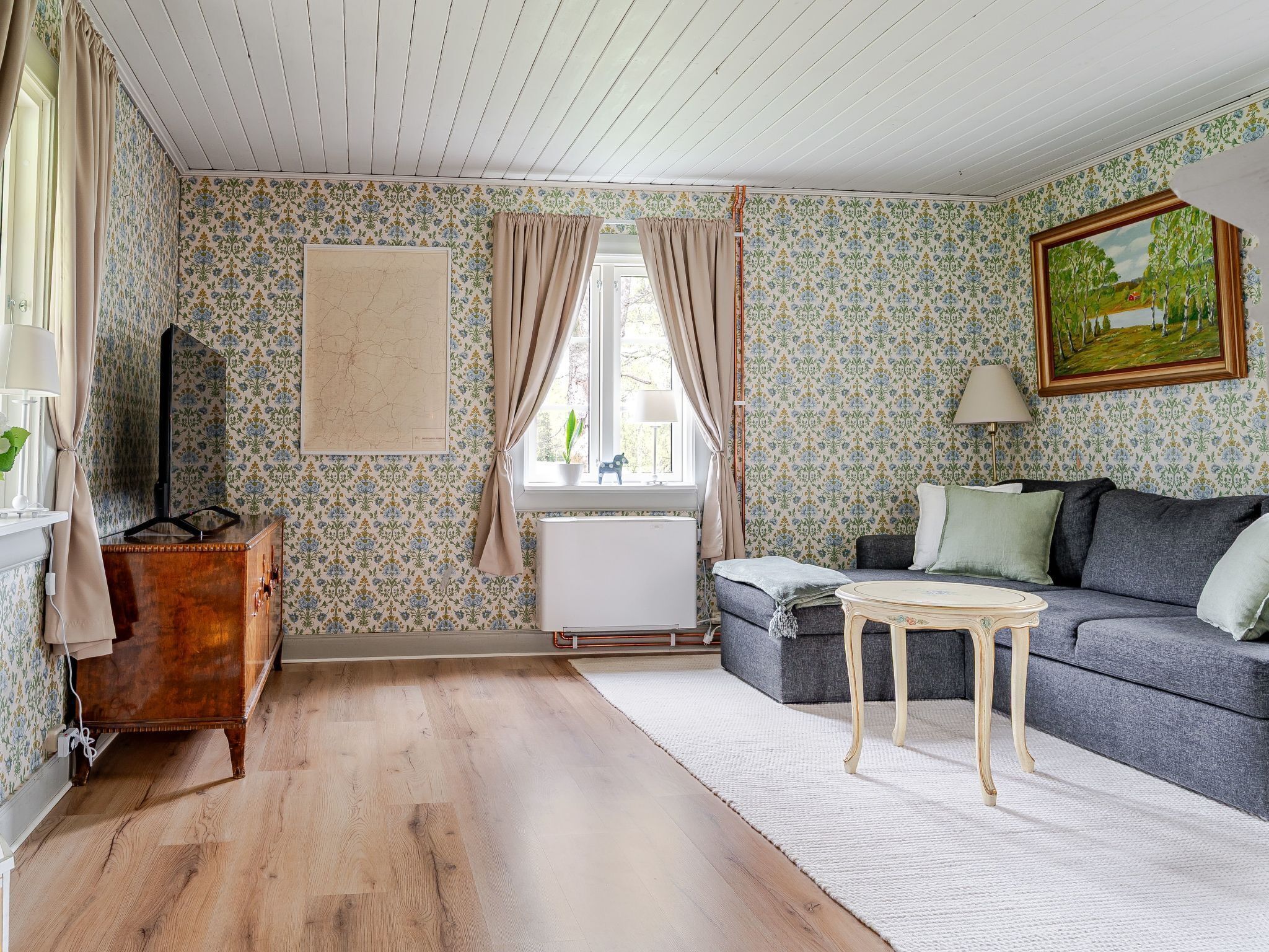 A cozy living room with patterned wallpaper, a wooden cabinet, a grey sofa, a small table, and a painting on the wall.