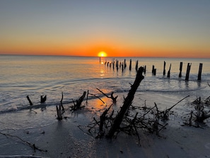 Een strand bij zonsondergang met houten palen in het water en drijfhout op het strand.