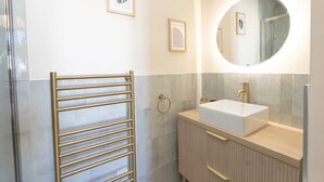 A modern bathroom with a glass shower enclosure, a gold towel rack, a white sink, and a mirror with a round frame. - Renovated apartment near the beach (Antibes)