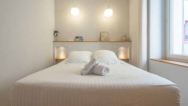 A neatly made bed with white linens and two pillows, a small shelf with decorative items, and wall-mounted lighting. - Luxurious Retreat in Old Antibes & beach (Antibes)