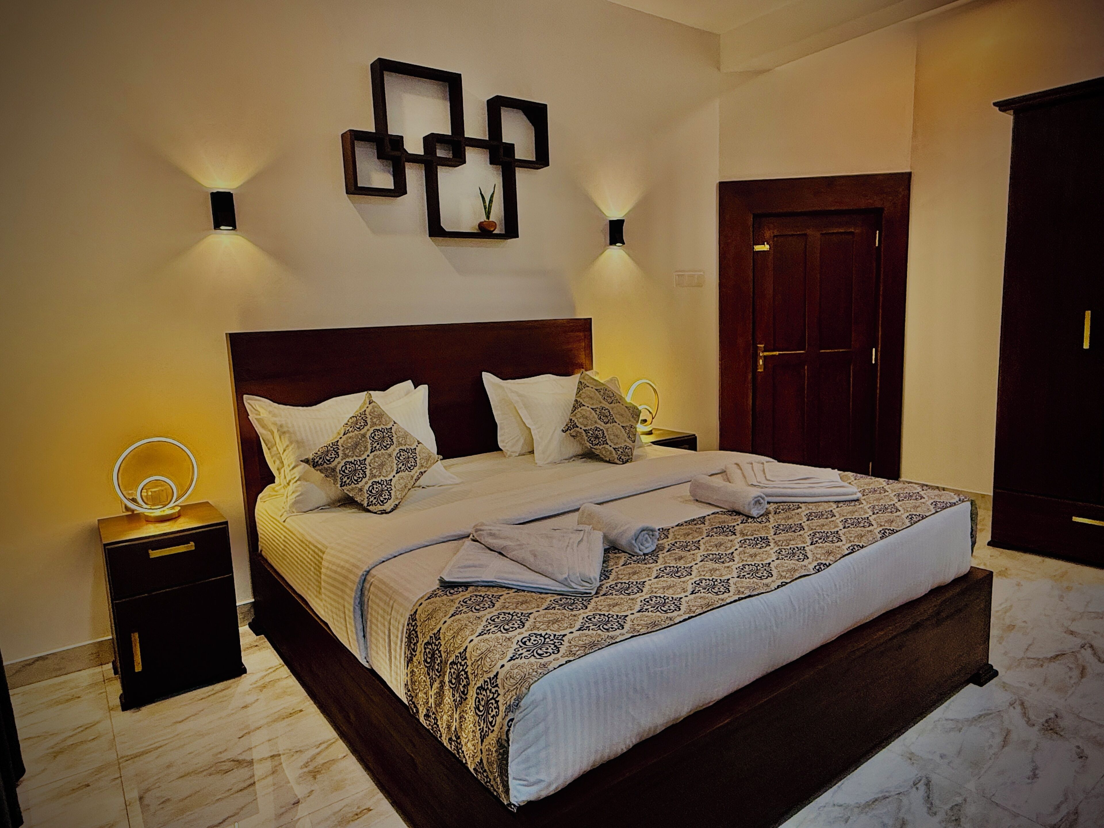 Una cama bien hecha con un edredón estampado, dos mesitas de noche con lámparas, una lámpara de pared y una puerta de madera.