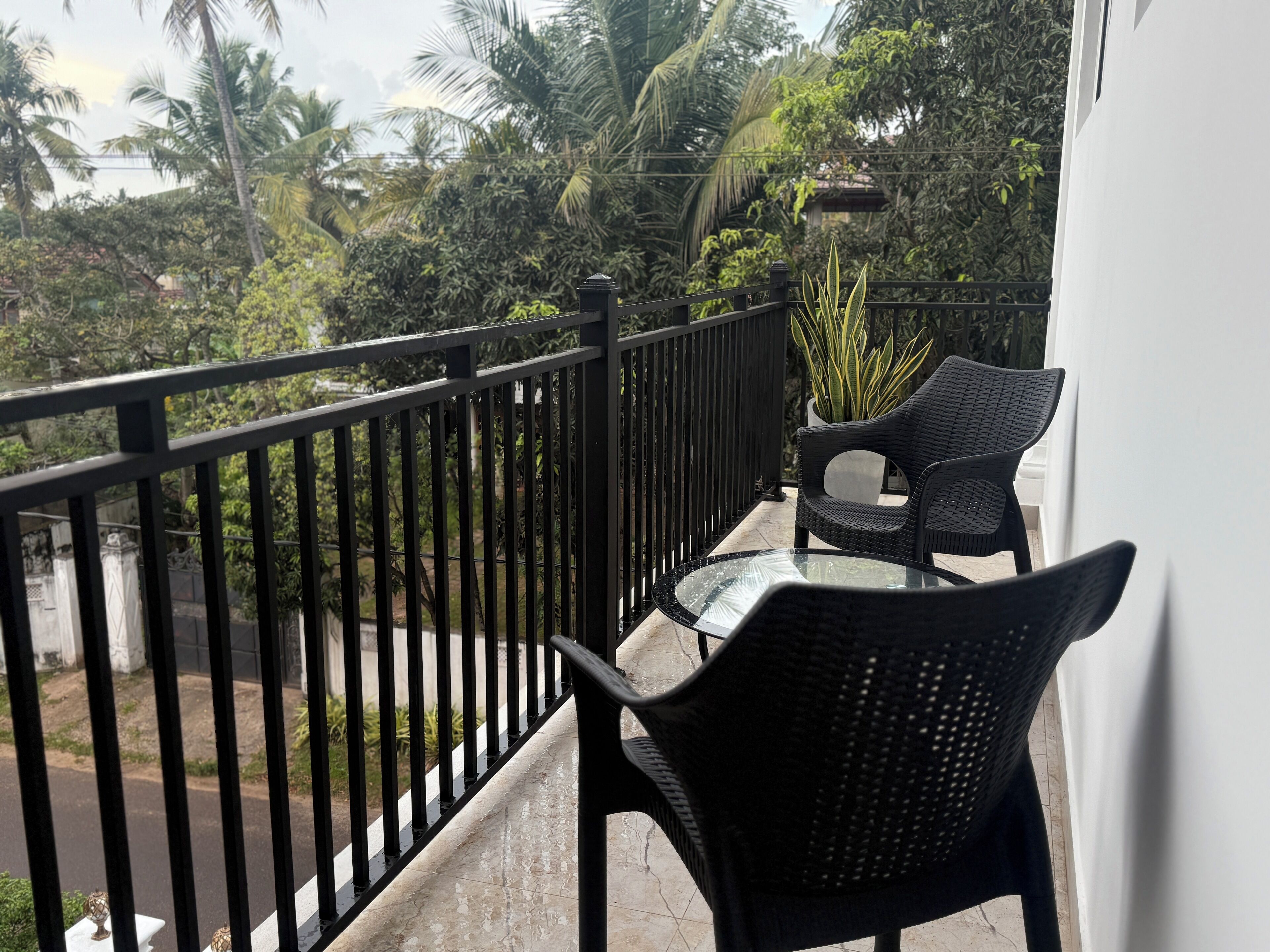 A balcony with a glass table and two wicker chairs, overlooking a garden with palm trees and a street.
