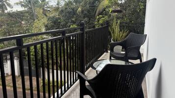 A balcony with a glass table and two wicker chairs, overlooking a garden with palm trees and a street.